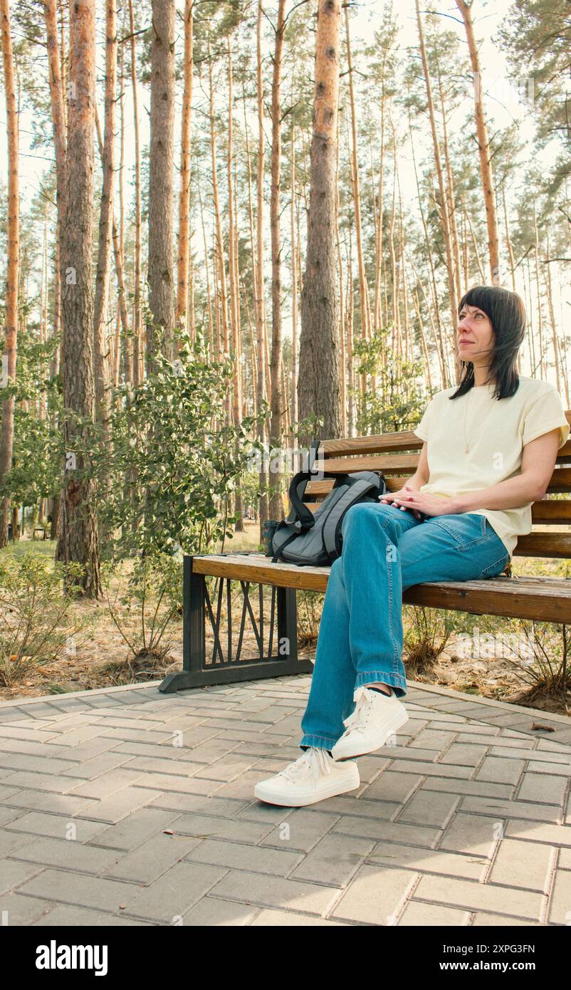 Jeune femme en vêtements décontractés assis sur le banc dans le parc. Fille en Jean bleu et T-shirt jaune se relaxant dans la forêt de pins. Tranquillité dans la vie Banque D'Images