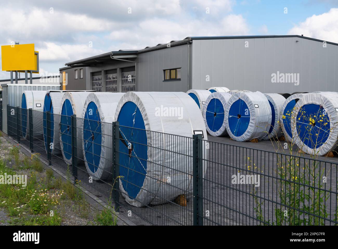 De grandes bobines bleues sont soigneusement disposées à l'extérieur d'une installation de stockage, avec une clôture séparant la zone de l'environnement environnant sous ciel nuageux. Banque D'Images