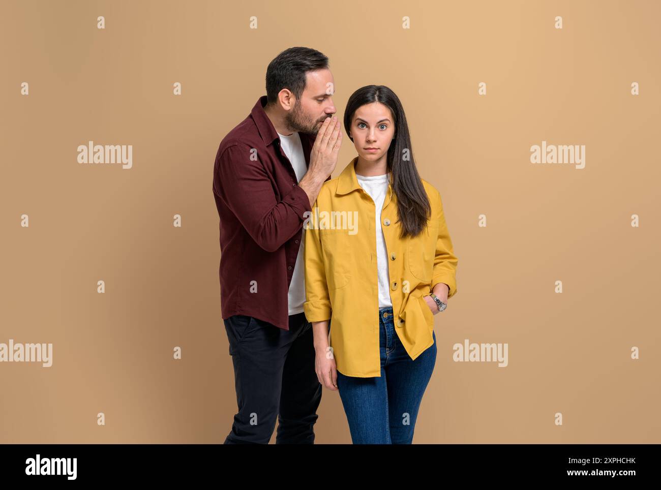 Petite amie choquée avec les sourcils levés écoutant son petit ami chuchoter secret dans ses oreilles. Jeune couple discutant de commérages privés en se tenant debout Banque D'Images