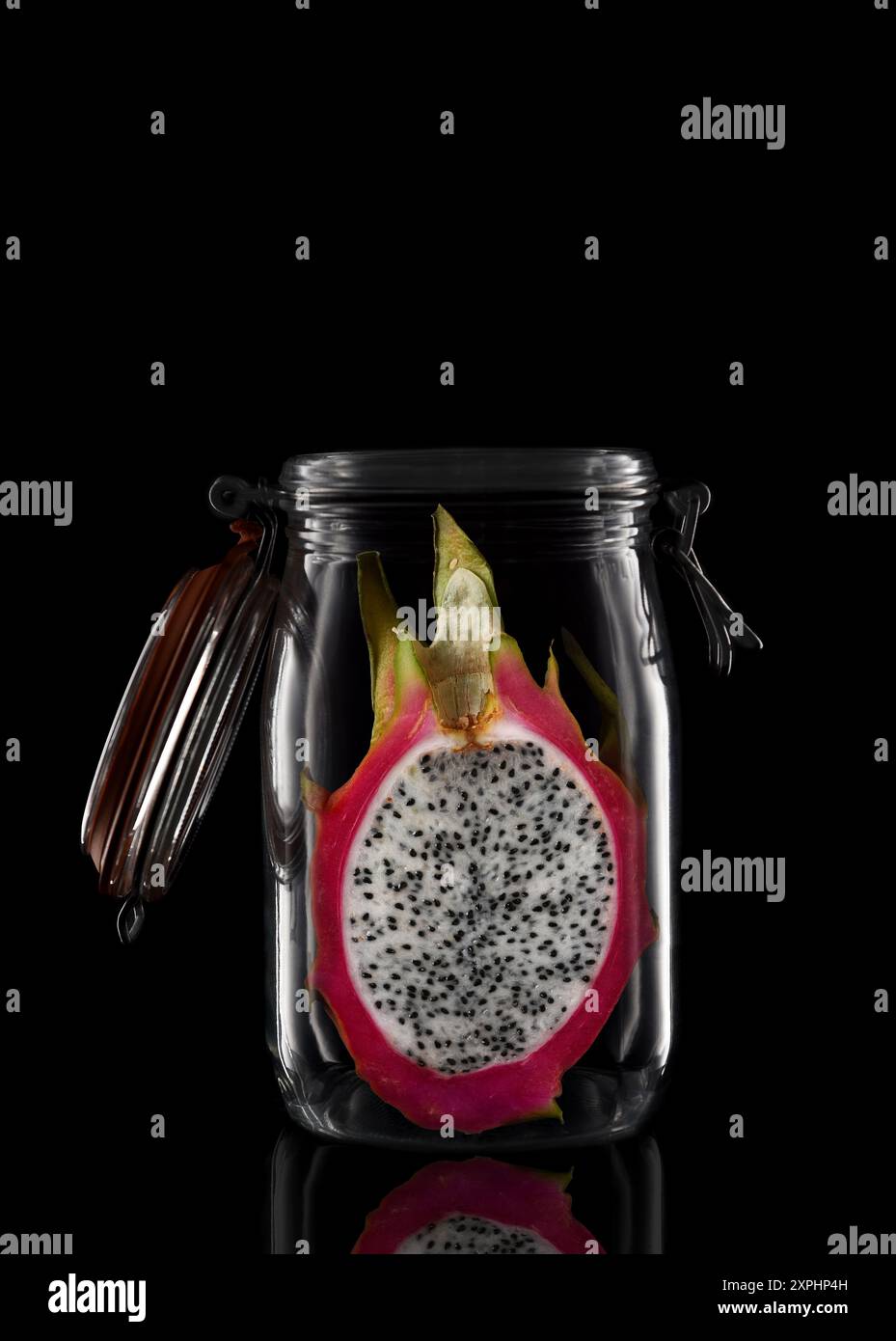 Un pot de stockage ou de mise en conserve en verre isolé sur noir avec réflexion, avec couvercle ouvert. Avec Un fruit de dragon coupé en deux Banque D'Images