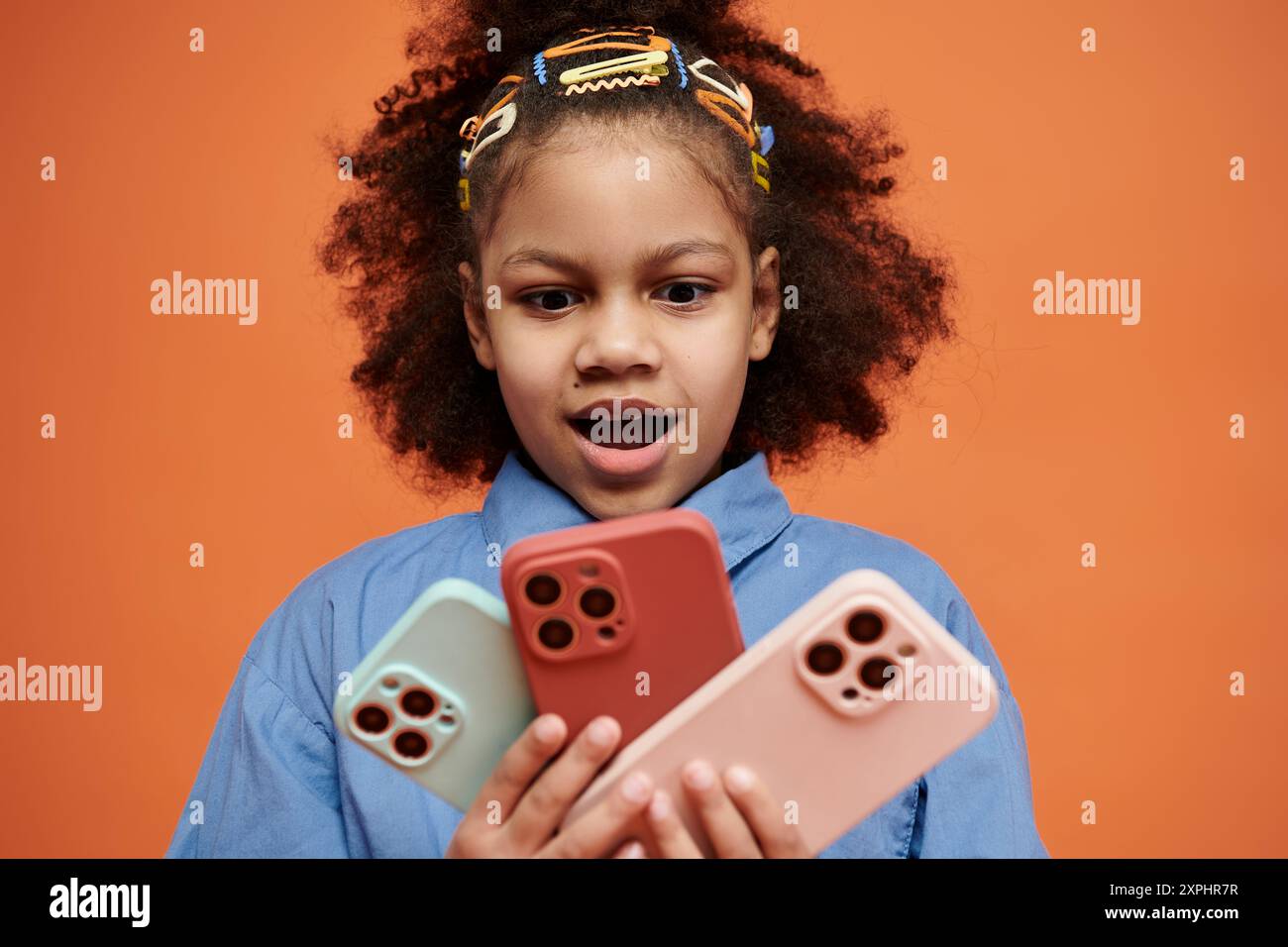 Une jeune fille afro-américaine dans une tenue élégante se tient devant un fond orange, tenant trois étuis de téléphone de couleur différente. Banque D'Images