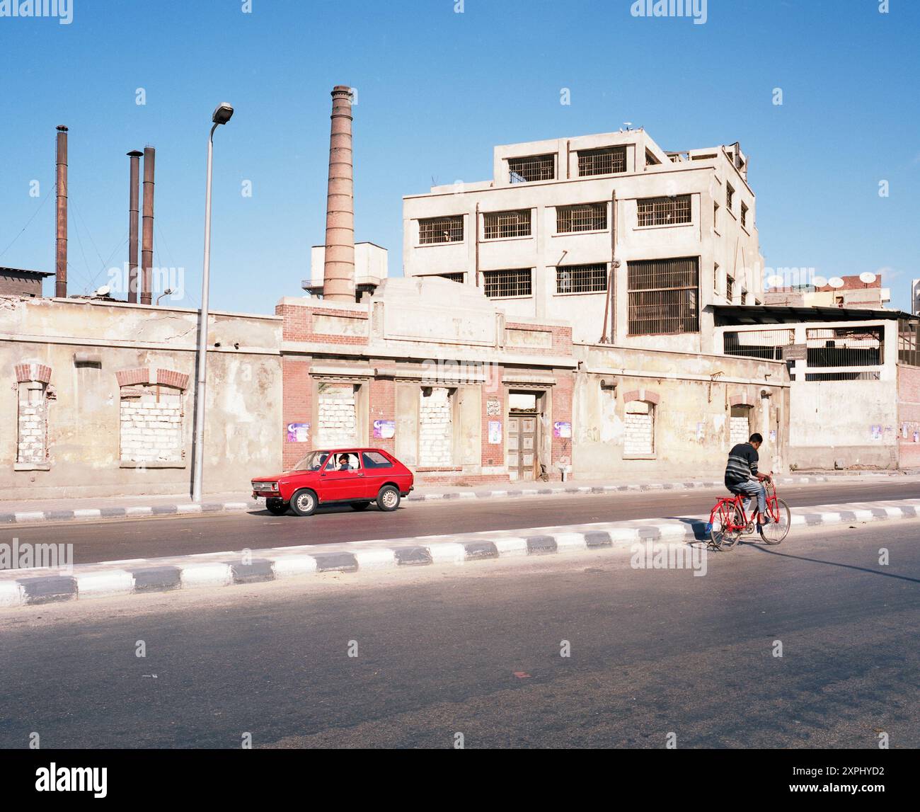 Une scène de rue au canal Mahmoudiya à Alexandrie, Egypte en 2006. L'image montre un bâtiment industriel, une voiture rouge et une personne sur un vélo, capturant la vie quotidienne et l'architecture. Banque D'Images