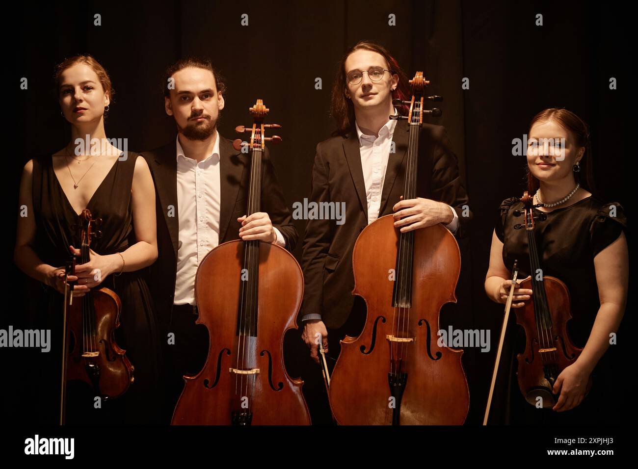 Portrait de groupe de classique de quatuor à cordes regardant la caméra tout en se tenant debout sur scène avec une lumière supérieure chaude contre des rideaux noirs Banque D'Images