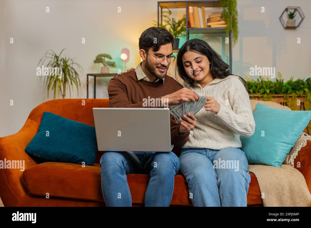 Heureux jeune couple arabe comptant de l'argent et des billets de réservation pour des vacances se trouve sur le canapé dans le salon à la maison. Famille diversifiée détenant des billets de banque planification de budget d'investissement en ligne Home Finance Banque D'Images
