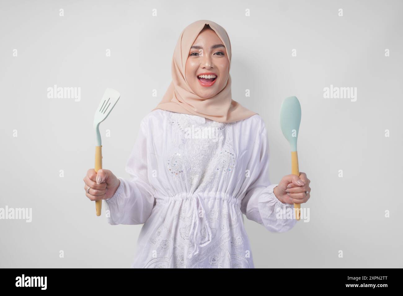 Portrait d'une femme musulmane asiatique heureuse en hijab voile tenant spatule et ustensiles de cuisine tout en souriant joyeusement. Concept de Ramadan Banque D'Images
