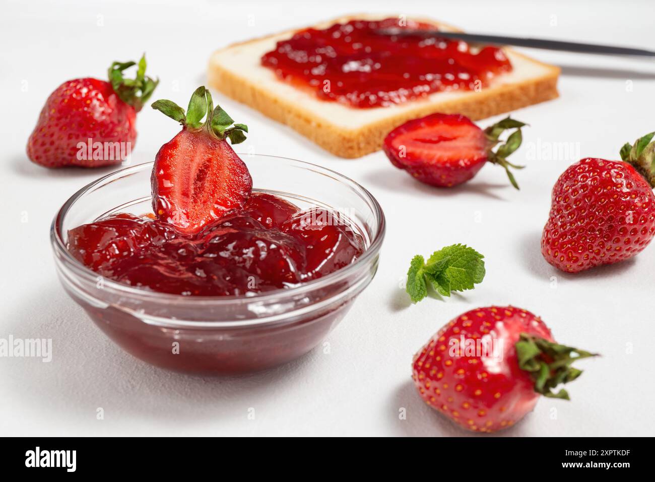 Un bol de confiture de fraises avec des fraises fraîches et une tranche de pain grillé étalé avec de la confiture, disposés sur un fond blanc Banque D'Images