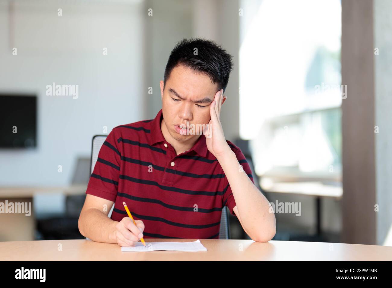 Un jeune adolescent fait un examen ou des devoirs dans une salle de classe, paraissant stressé tout en écrivant avec un crayon sur le bureau Banque D'Images