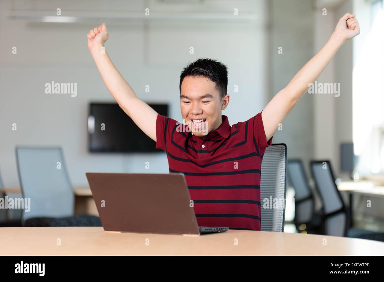 Un jeune étudiant est assis à une table avec les bras levés en l'air en célébration devant un ordinateur portable sur la table Banque D'Images