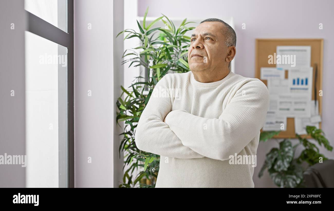 Un homme hispanique contemplatif d'âge moyen se tient debout les bras croisés dans un cadre de bureau moderne. Banque D'Images