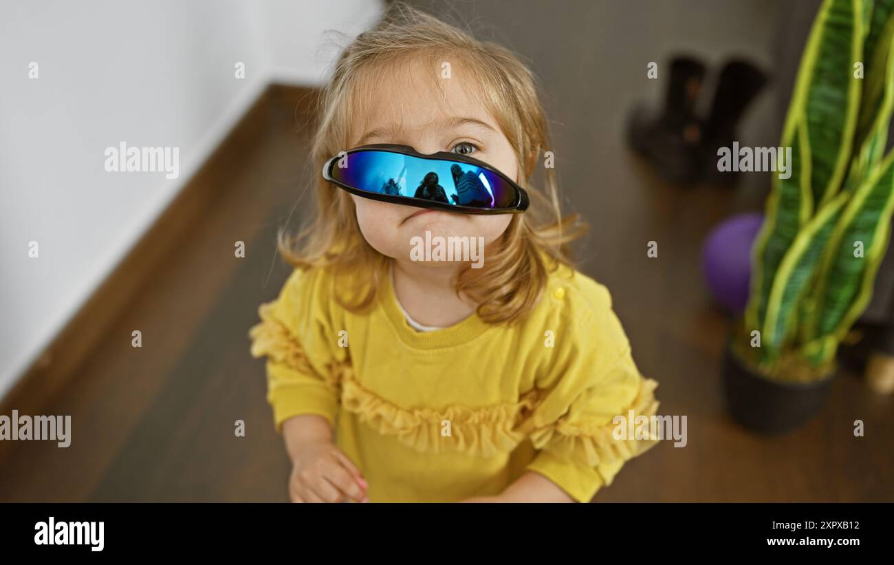 Adorable fille blonde caucasienne portant des lunettes de soleil surdimensionnées à l'intérieur, montrant la méchanceté de l'enfance dans un cadre de maison. Banque D'Images