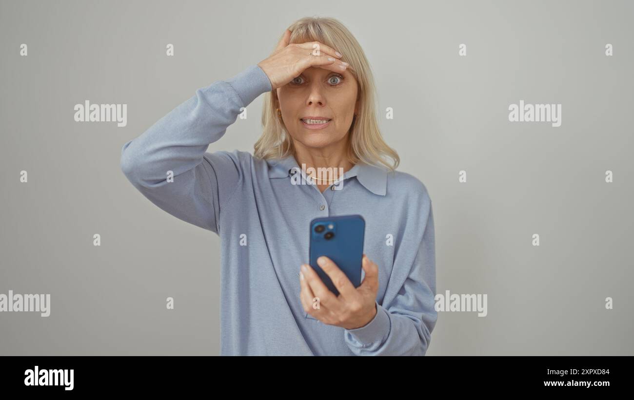 Une femme blonde surprise tient un smartphone sur un fond blanc, exprimant le choc ou la confusion. Banque D'Images