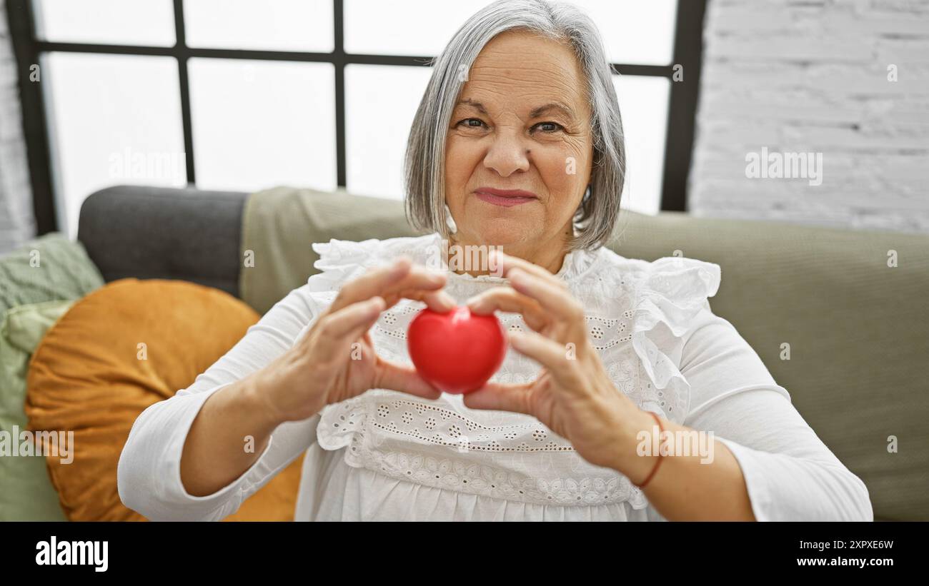 Une femme âgée souriante tient une forme de cœur rouge à la maison, représentant la santé ou l'amour dans un cadre confortable de salon. Banque D'Images