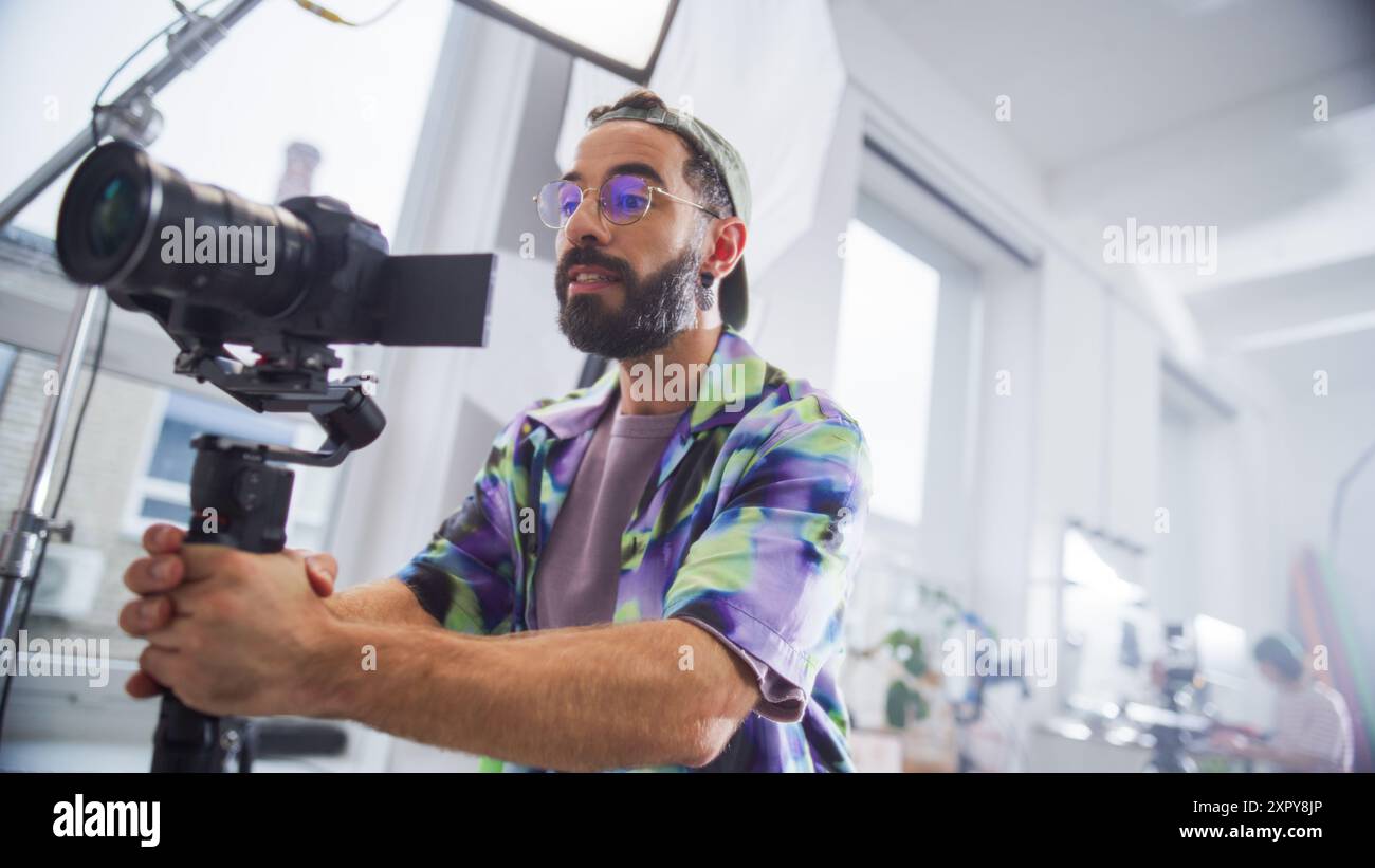 Directeur de la photographie masculin concentré avec barbe, portant une chemise colorée, utilise un appareil photo professionnel dans un cadre de studio lumineux, capturant du contenu attrayant pour un film ou un projet publicitaire. Banque D'Images