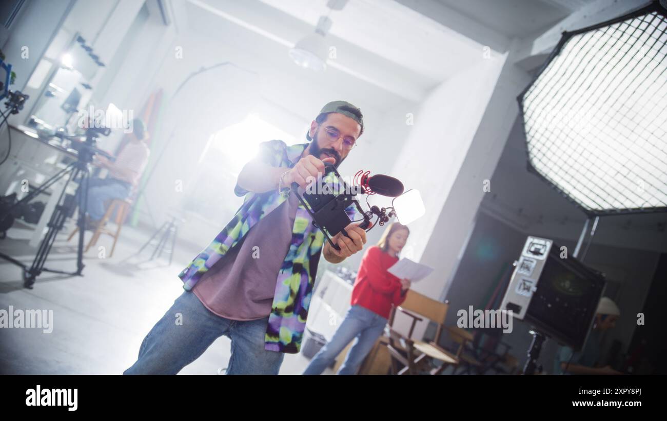Jeune cinéaste masculin concentré avec barbe, portant Une casquette et une chemise à carreaux, filmant activement dans Un cadre de studio professionnel, entouré d'équipements de tournage modernes et de membres de l'équipe. Banque D'Images