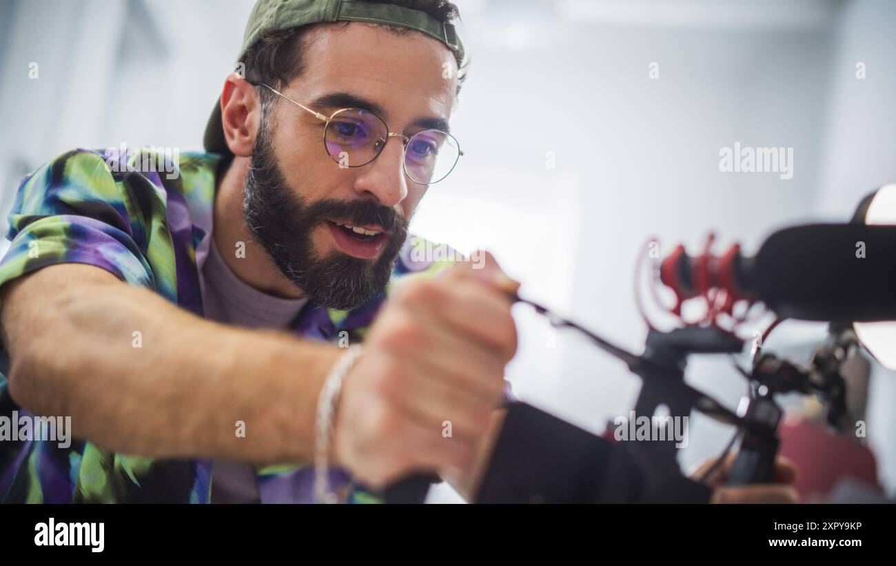 Un jeune cinéaste masculin concentré avec barbe et lunettes ajuste l'appareil photo lors d'un tournage en intérieur, capturant l'essence du cinéma moderne et de la publicité créative dans Un cadre professionnel. Banque D'Images