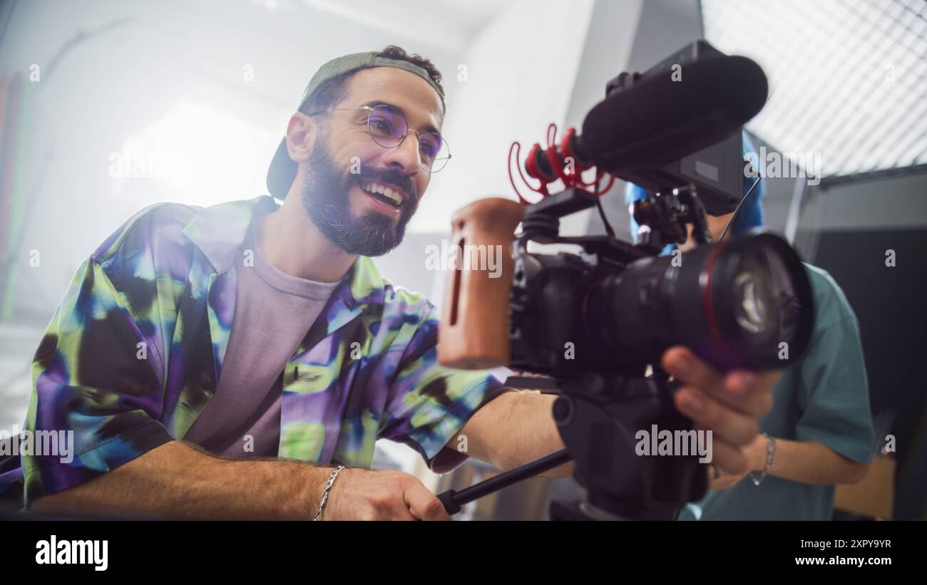 Jeune cinéaste masculin avec barbe et lunettes, souriant tout en utilisant Un appareil photo professionnel dans Un cadre de studio, capturant des séquences pour Un projet de publicité créative. Banque D'Images