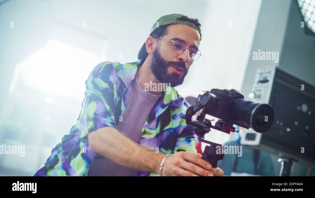 Jeune cinéaste masculin concentré avec barbe, réglage des paramètres sur Un appareil photo moderne lors d'un tournage en intérieur, port d'Une veste et de lunettes colorées. Créativité et professionnalisme dans la réalisation de films. Banque D'Images