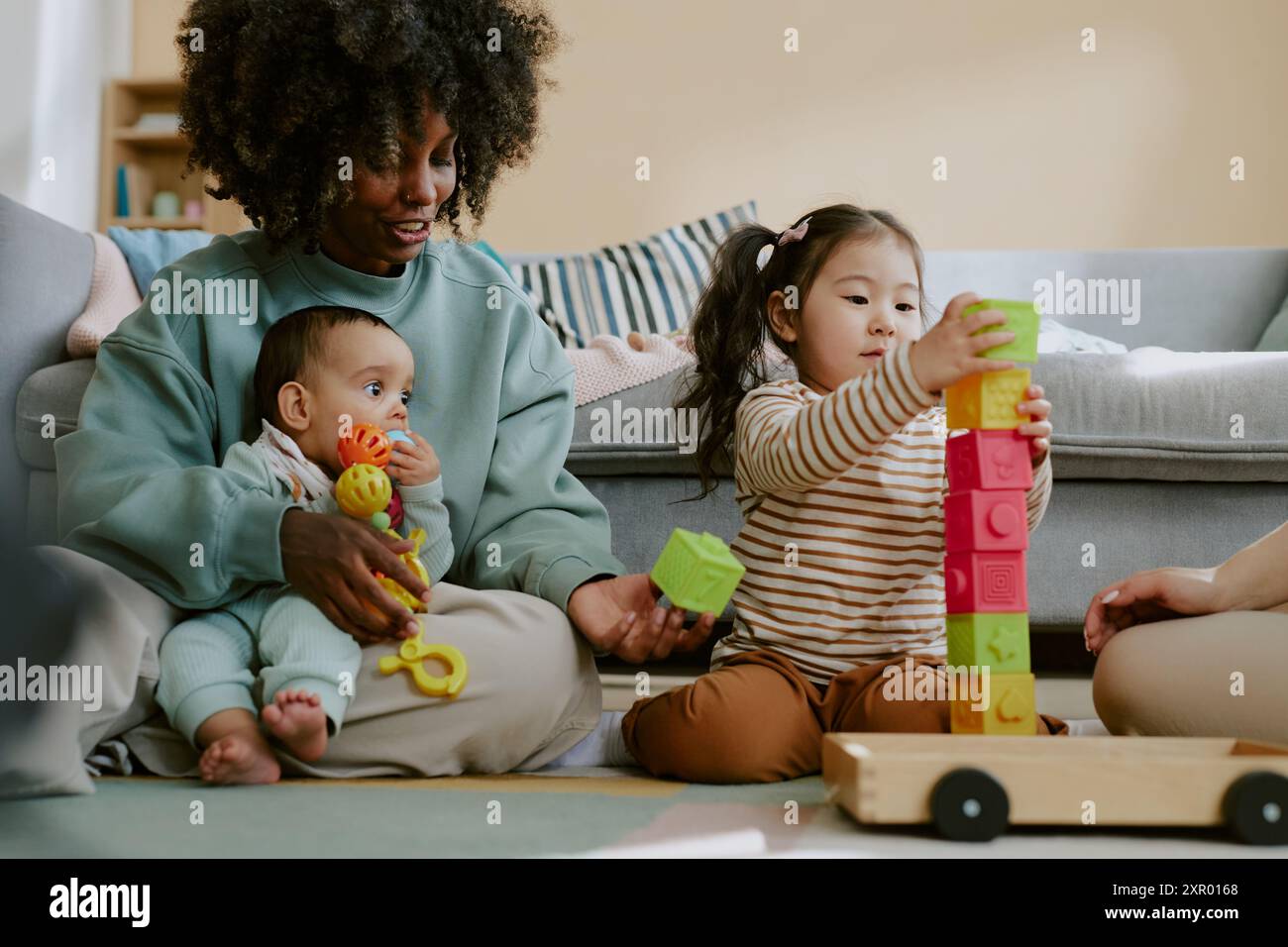 Maman afro-américaine tenant son bébé fils tout en aidant le tout-petit asiatique à construire une tour de briques Banque D'Images