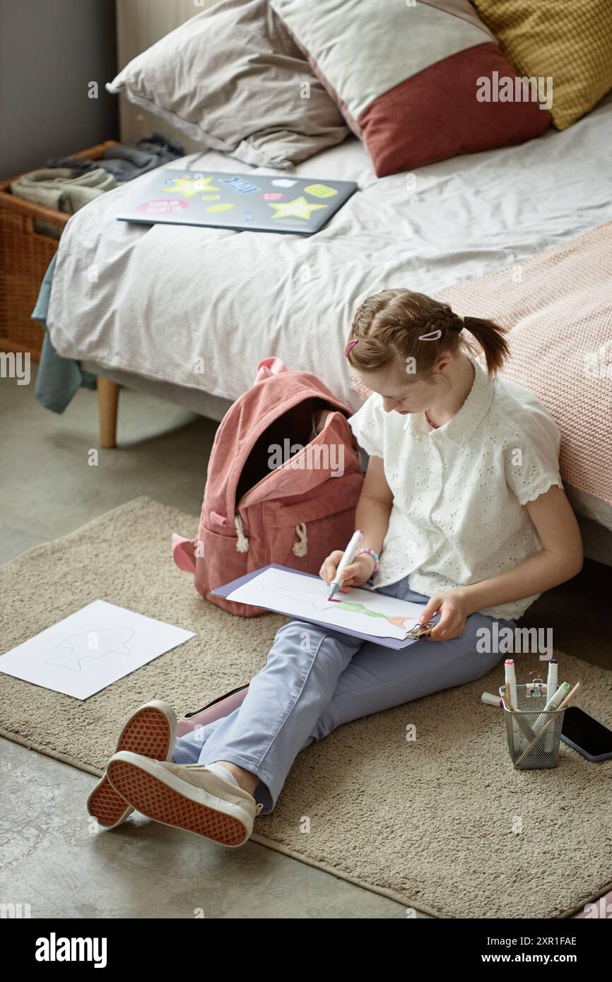 Jeune fille assise sur le sol de la chambre à coucher faisant des devoirs d'art avec sac à dos et matériaux de dessin, montrant la créativité et la concentration, atmosphère détendue et confortable Banque D'Images