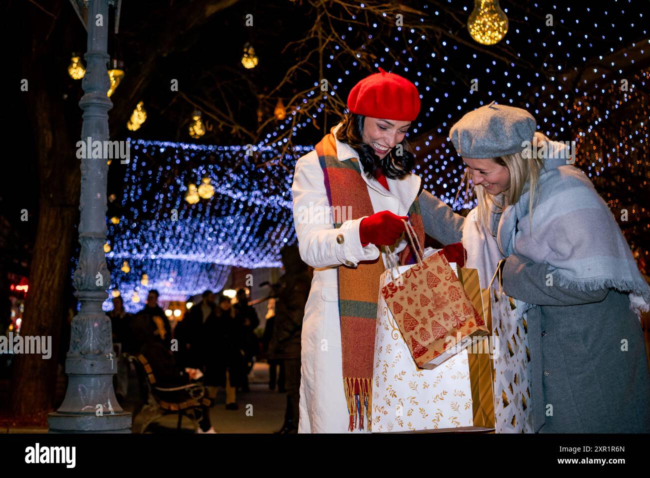 Les copines en manteau et bérets français avec sacs à provisions apprécient les joyeux achats de Noël sur une place de la ville décorée de façon festive. Banque D'Images