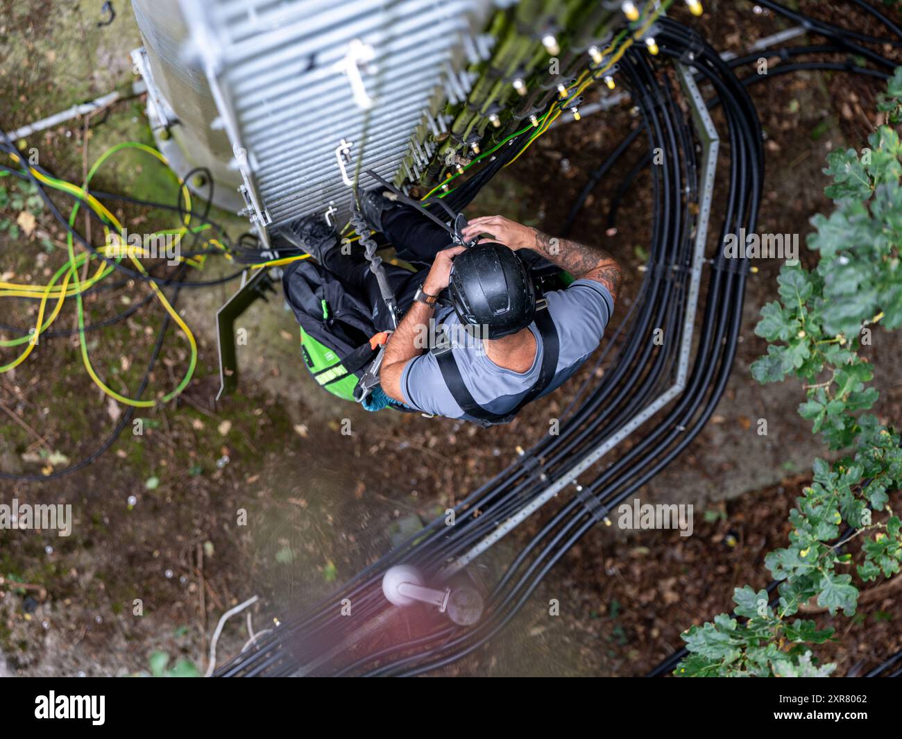 Technicien en télécommunications travaille en hauteur, portant un harnais de sécurité et un casque, installant des câbles à fibres optiques sur une structure métallique Banque D'Images