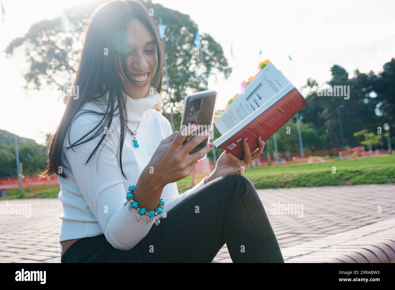 Heureuse jeune femme latine à l'extérieur lisant des messages sur son téléphone portable, elle est assise souriante en regardant le téléphone tout en tenant un livre avec l'autre Banque D'Images