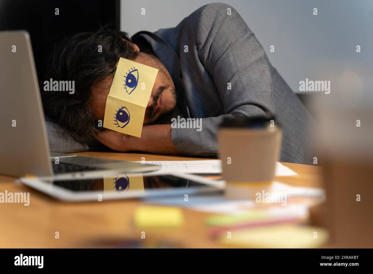 Homme adulte joyeux se reposant dans le bureau avec des notes collantes sur les yeux. Homme endormi ayant des autocollants avec les yeux peints sur le visage se relaxant avec les mains derrière Banque D'Images