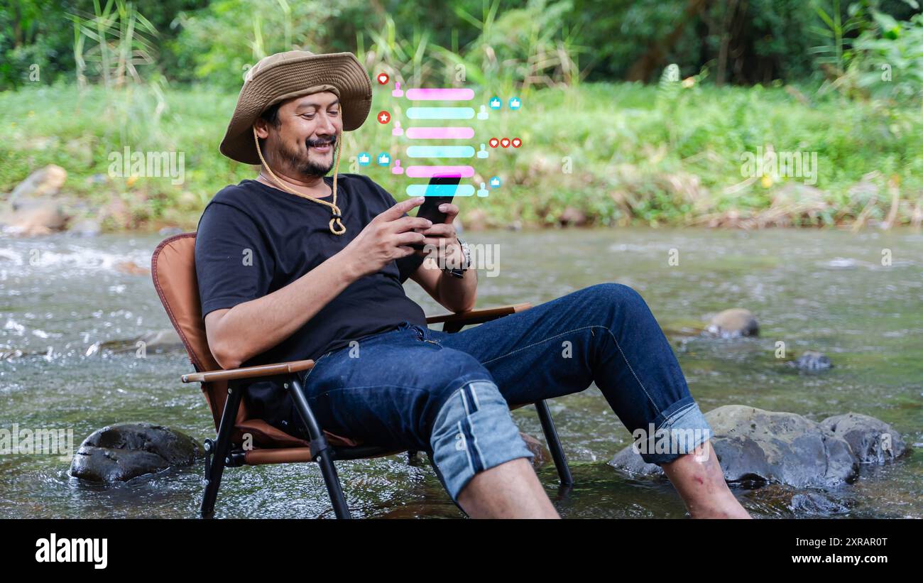 Un bel homme avec une barbe utilise un smartphone assis sur une chaise, libérant une image holographique d'icônes liées aux médias sociaux sur un lac de montagne. Banque D'Images