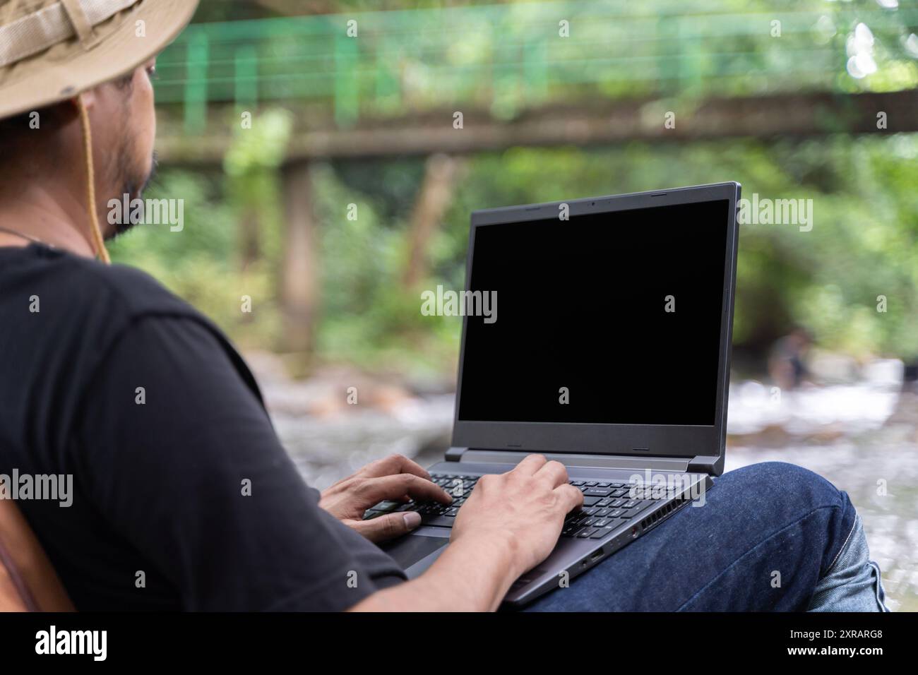 Beau homme asiatique de travail barbu assis sur la chaise de camping et travaillant sur l'écran blanc noir d'ordinateur portable près du ruisseau au milieu du parc dans la forêt extérieure A. Banque D'Images