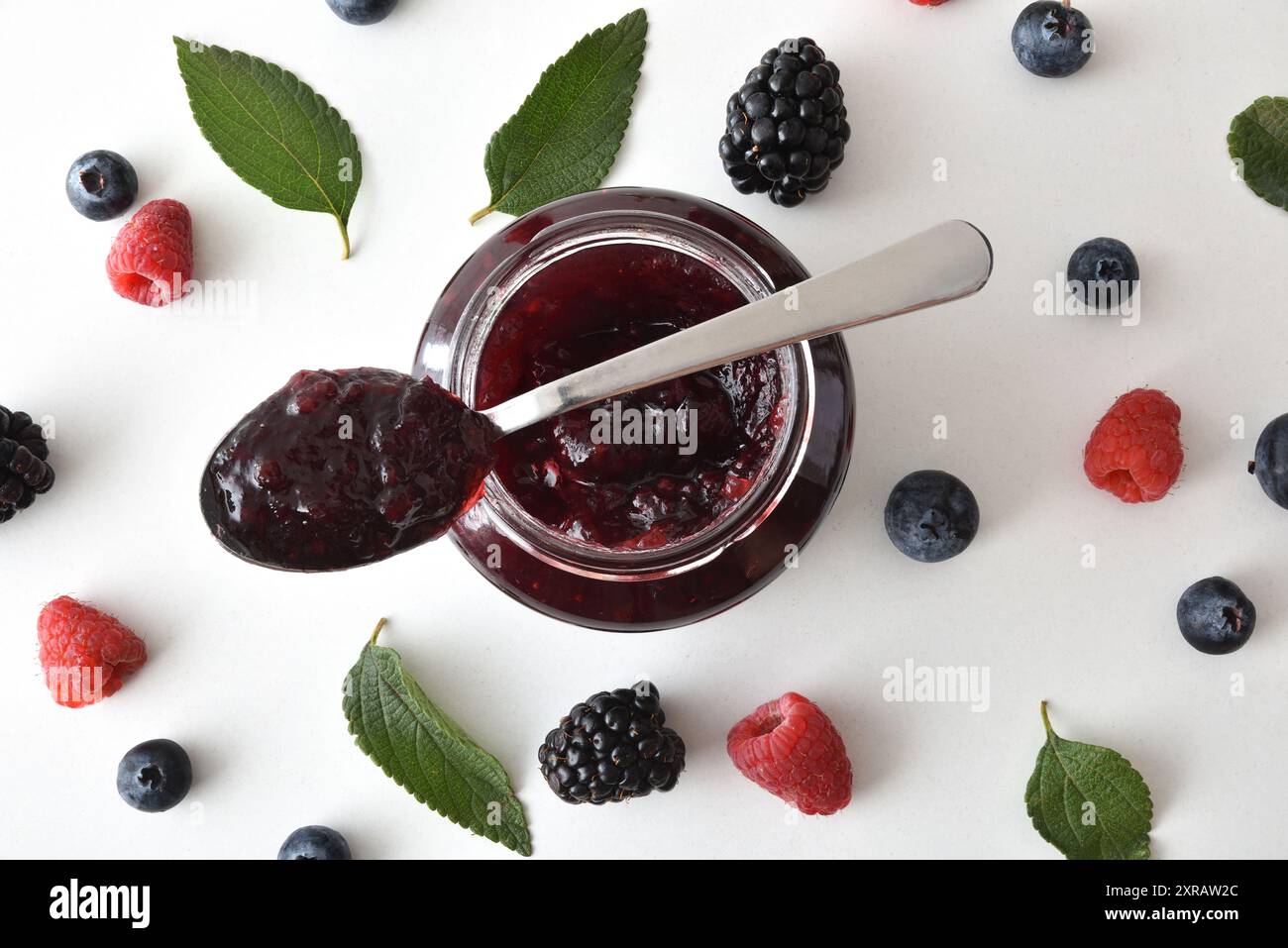 Fond avec cuillère sur pot avec de la confiture de baies et des mûres, des myrtilles et des framboises sur une table blanche avec des feuilles. Vue de dessus. Banque D'Images