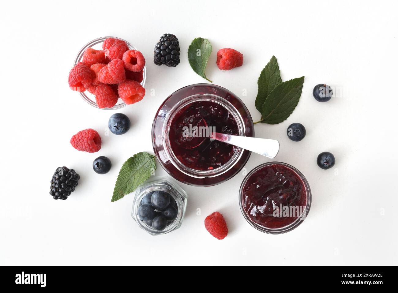 Fond avec cuillère sur pot avec de la confiture de baies et des mûres, des myrtilles et des framboises sur une table blanche avec des feuilles. Vue de dessus. Banque D'Images