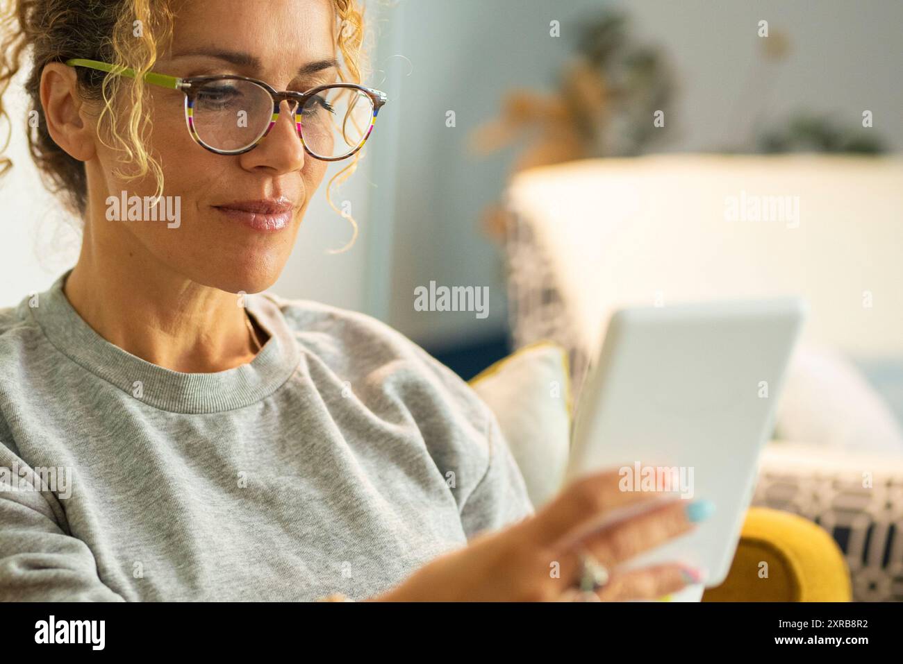 Gros plan potrait de femme moderne femmes adultes lisant le livre à partir de la tablette de lecteur électronique. Dame portant des lunettes et sourire appréciant le temps de détente à la maison assis sur le canapé Banque D'Images