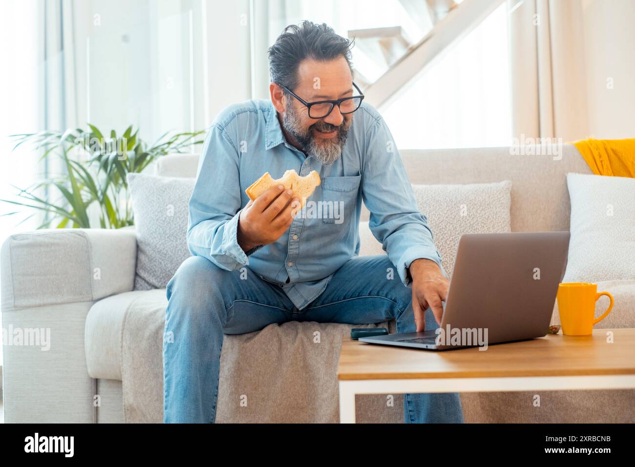 Un homme dans des vêtements décontractés en utilisant un ordinateur portable à la maison assis sur un canapé et manger sandwich souriant. Les gens avec de la nourriture et de la technologie. Activités de loisirs en intérieur. Informatique et nutrition. Navigation sur la connexion Internet Banque D'Images