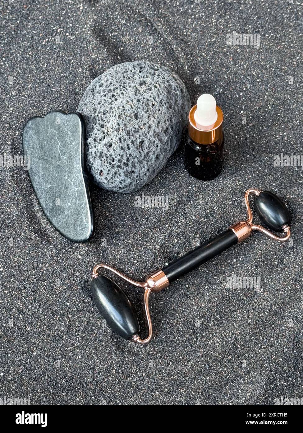 Outils de soin de la peau sur sable noir avec pierre de gouache, bouteille compte-gouttes et rouleau facial Banque D'Images