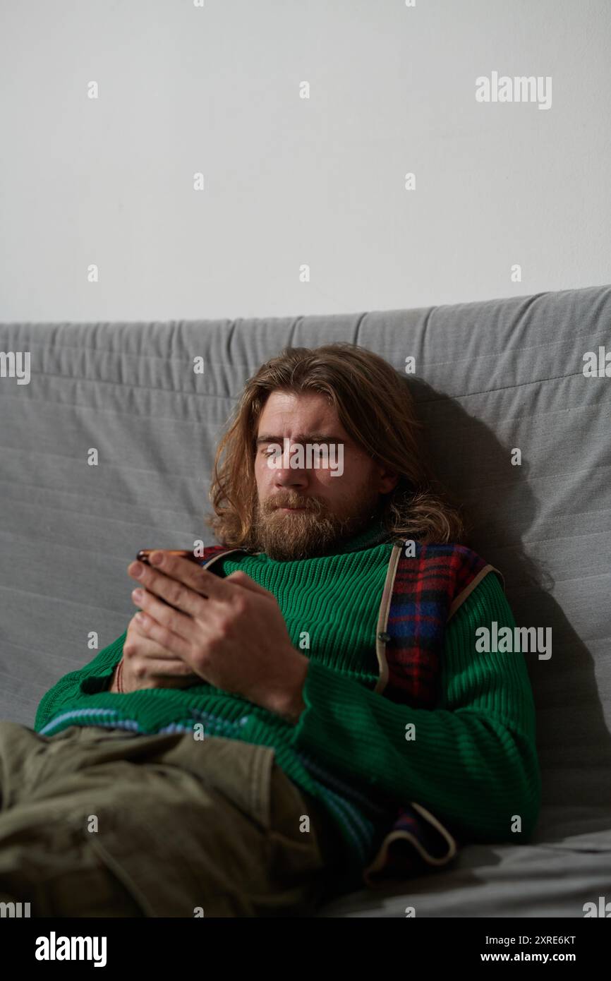 Homme barbu portant un pull vert et une écharpe à carreaux assis sur le canapé et tapant sur le téléphone, affichant une expression focalisée sans regarder la caméra Banque D'Images