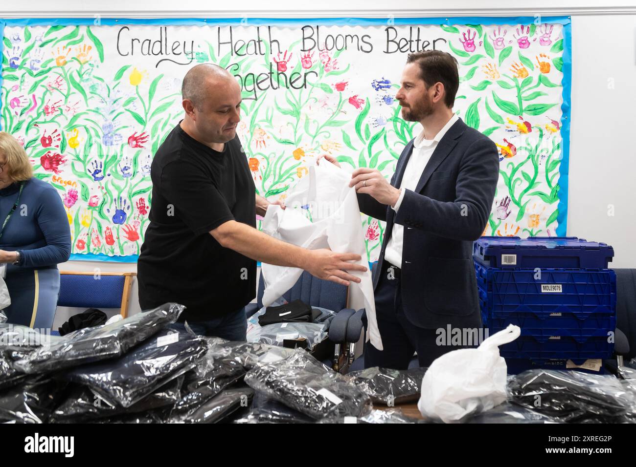 Cradley Heath, West Midlands, Royaume-Uni. Samedi 10 août 2024. Alex Ballinger, député travailliste de Halesowen dans le Black Country, aide Adam Banks, bénévole communautaire, à distribuer un uniforme scolaire d'occasion dans un centre communautaire de Cradley Heath, une région économiquement en difficulté dans le Black Country, dans les West Midlands. Des milliers d'uniformes scolaires locaux sont donnés, lavés, emballés et remis à la population locale pour les réutiliser pour aider à faire face à la crise du coût de la vie. Crédit : Peter Lopeman/Alamy Live News Banque D'Images