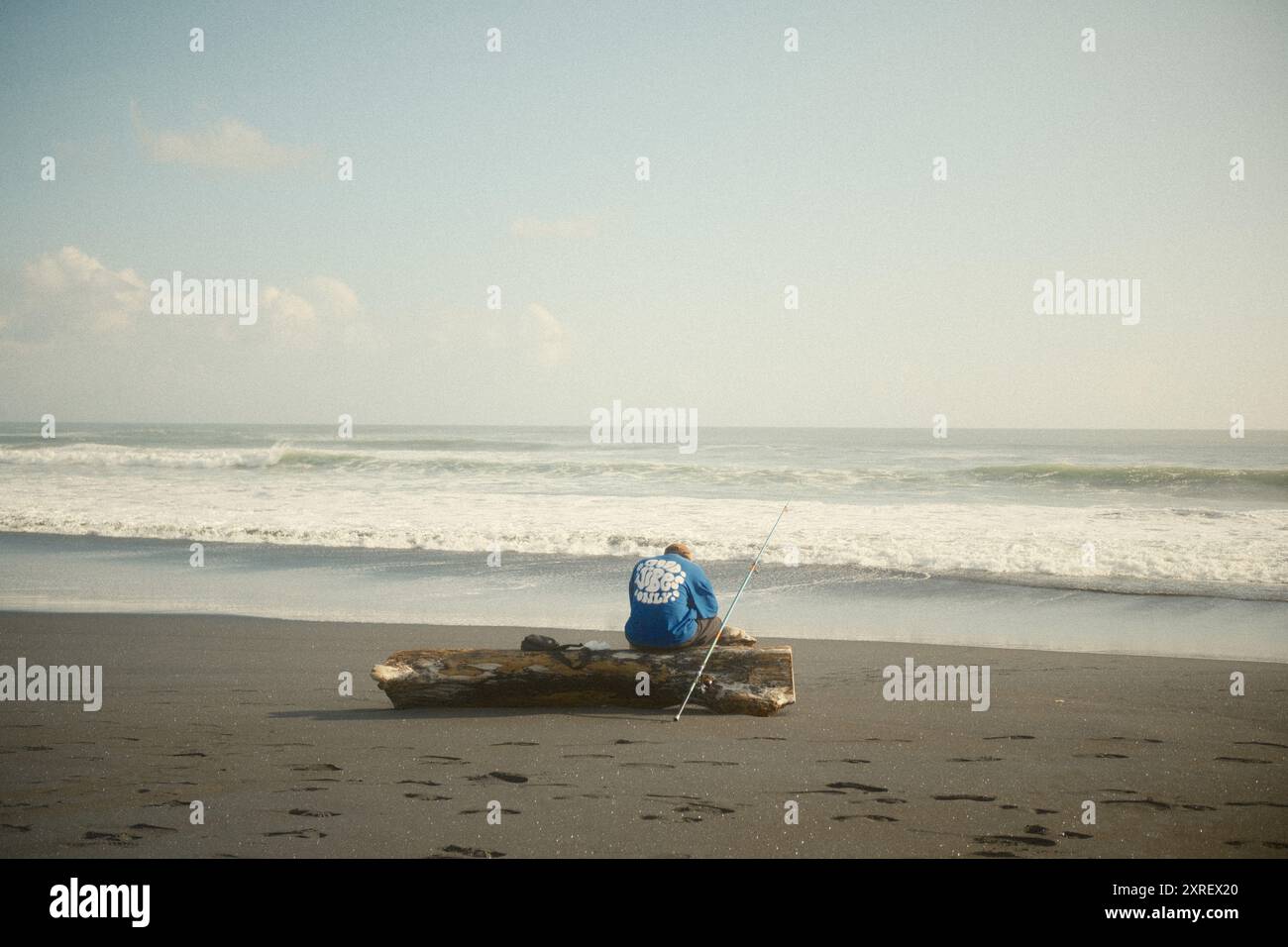 Tabanan, Bali, Indonésie - 9 août 2024 : un homme pêche ou fait du surf assis sur une bûche sèche au bord d'une plage de sable noir Banque D'Images