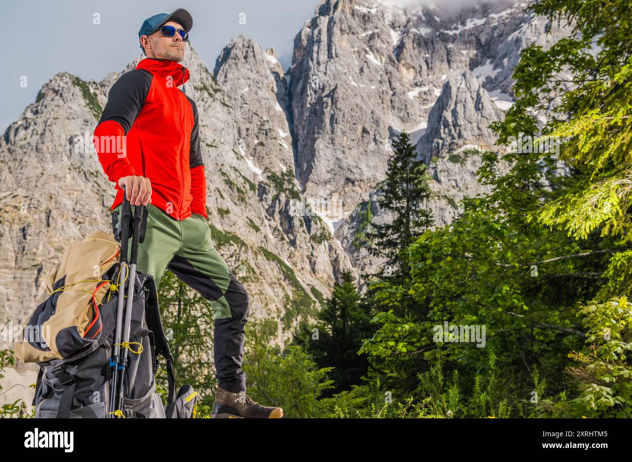 Un randonneur en veste rouge se tient confiant sur un affleurement rocheux, entouré de montagnes imposantes et d'arbres luxuriants. Banque D'Images