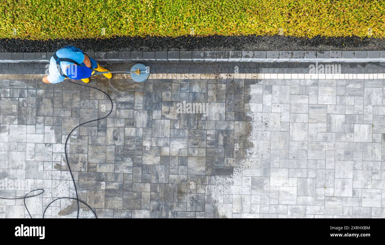 Un travailleur utilise une laveuse haute pression pour nettoyer un patio en pierre, révélant une surface fraîche et bien rangée dans un espace extérieur bien entretenu. Banque D'Images