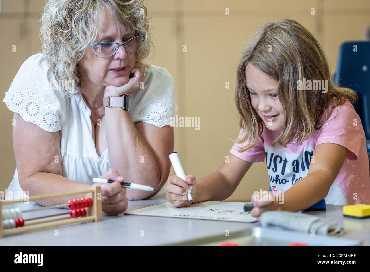 Un professeur aide une jeune fille avec ses devoirs de maths. La fille porte une chemise rose et utilise un marqueur blanc pour écrire sur un petit tableau blanc Banque D'Images