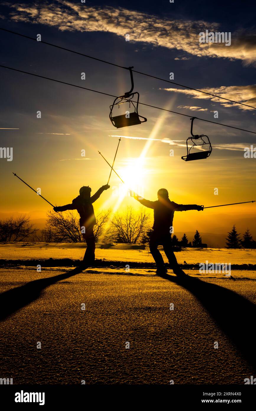 Deux skieurs célèbrent et profitent du coucher de soleil sur une montagne enneigée, avec des chaises de téléski au-dessus de la tête et des arbres au loin. Banque D'Images