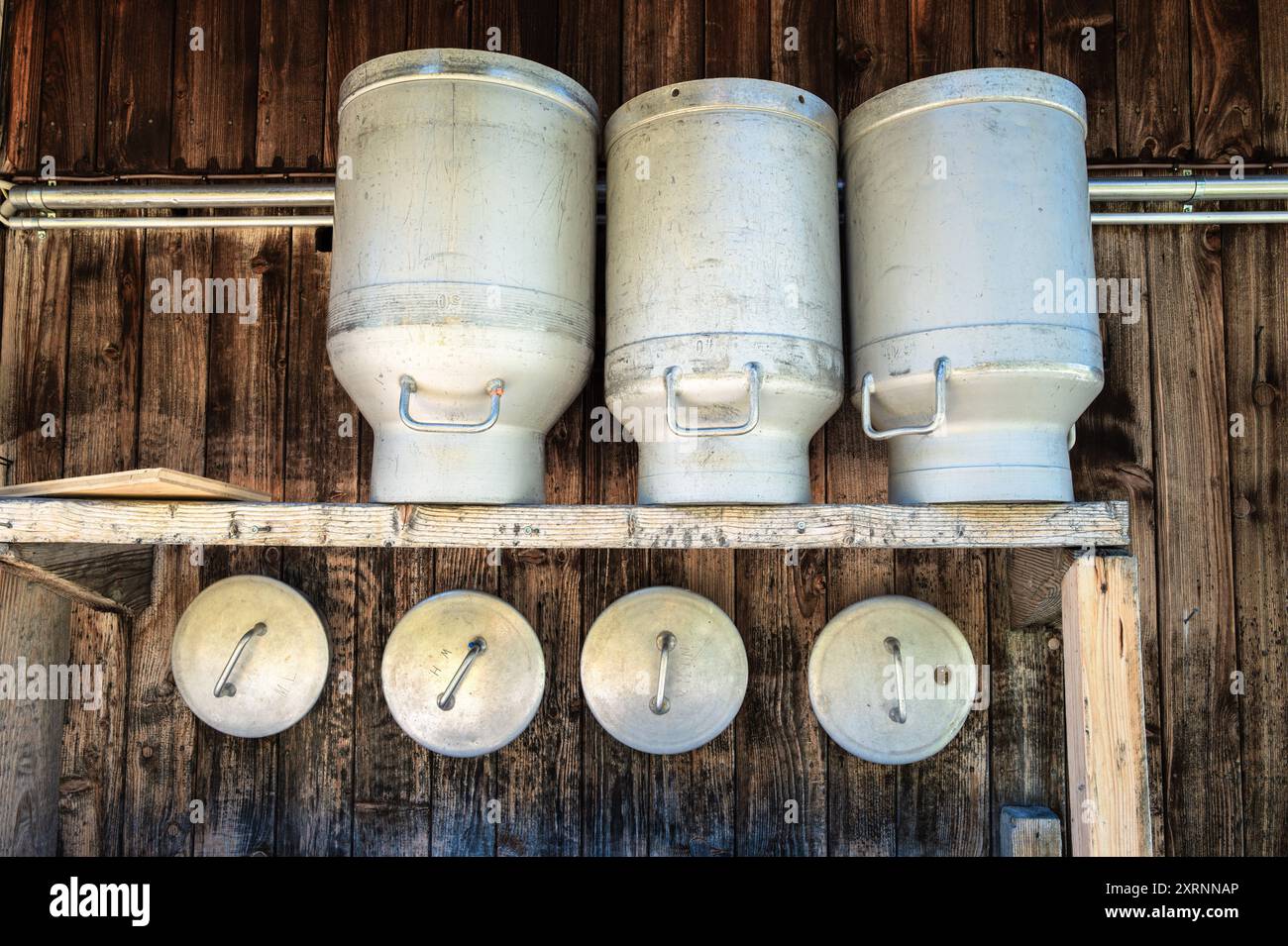 Conteneurs de lait en métal empilés et couvercles accrochés sur le mur en bois du chalet alpin. Banque D'Images