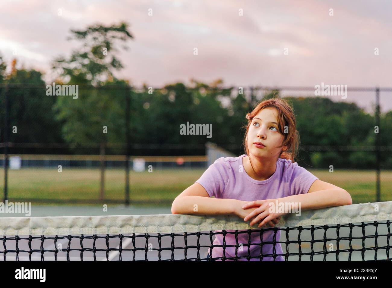 Une fille regarde penchée sur le filet au court de tennis au coucher du soleil levant les yeux Banque D'Images