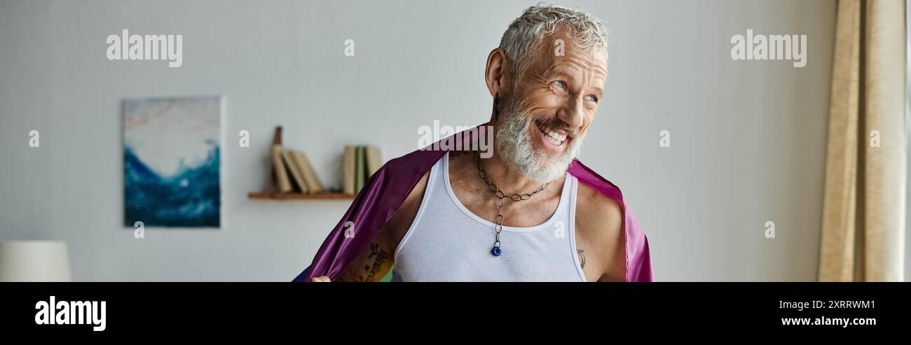 Un homme gay mature souriant avec des tatouages et des cheveux gris tient un drapeau arc-en-ciel tout en se tenant debout dans sa maison. Banque D'Images