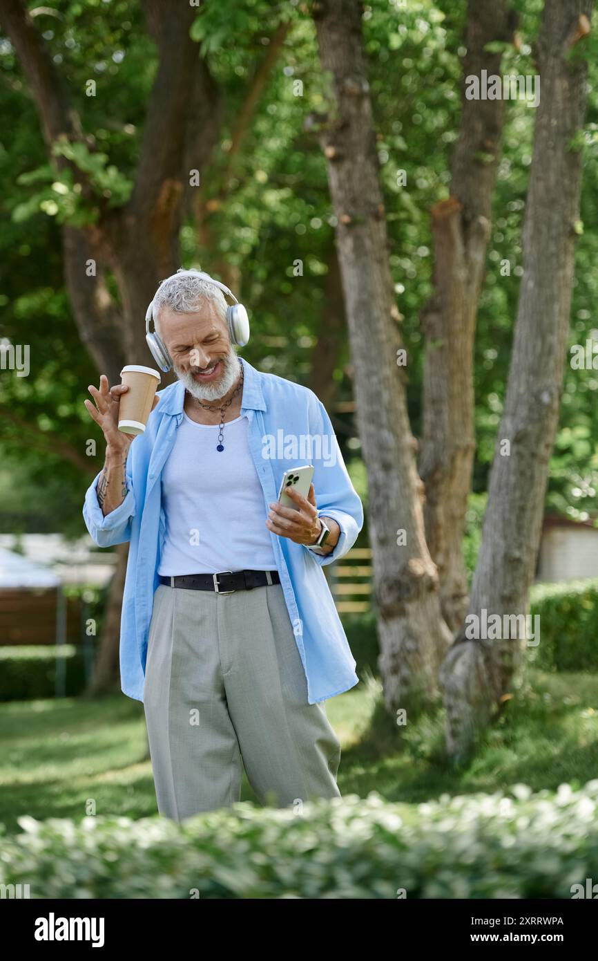 Homme avec des tatouages, barbe grise profite d'une journée ensoleillée dans le parc, écoutant de la musique avec du café. Banque D'Images