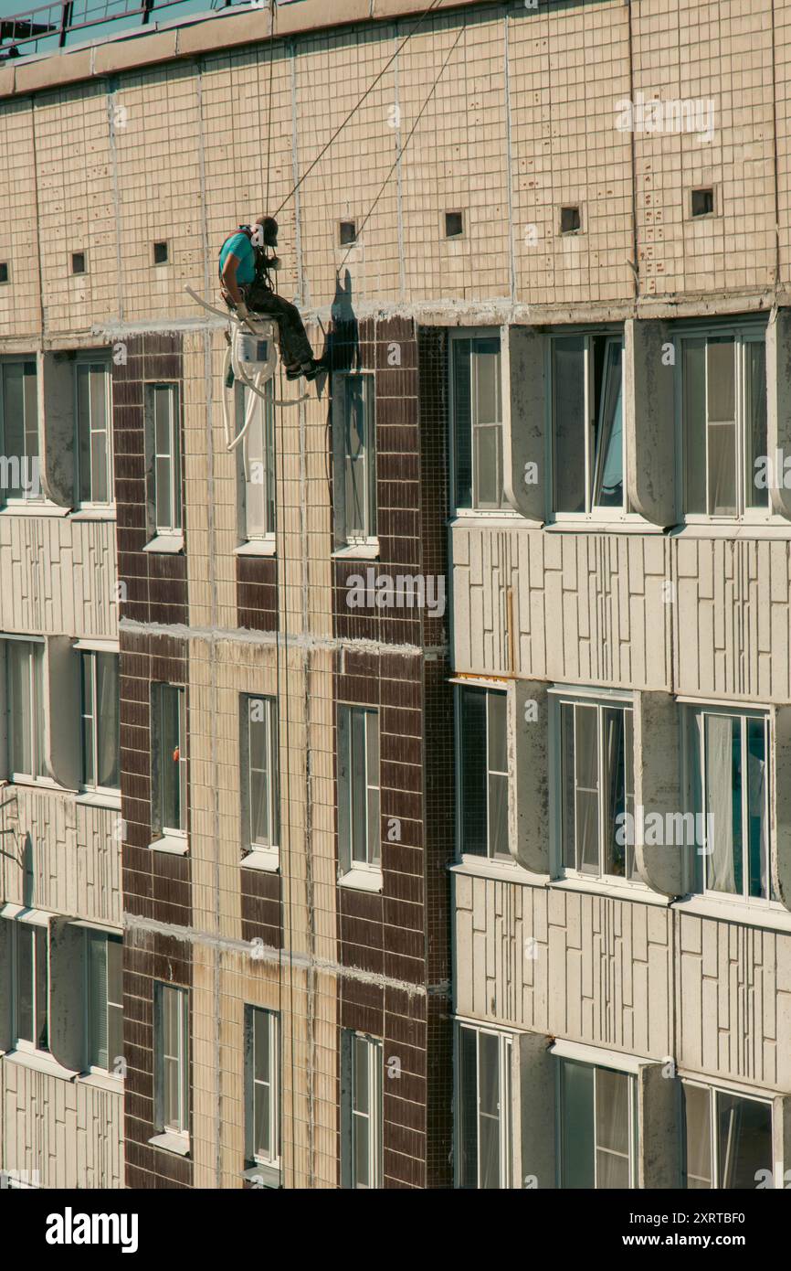 Un grimpeur industriel qualifié est vu réparer des fissures à l'extérieur d'un immeuble de grande hauteur à l'aide d'un équipement spécialisé et de harnais de sécurité sur une surface brillante Banque D'Images