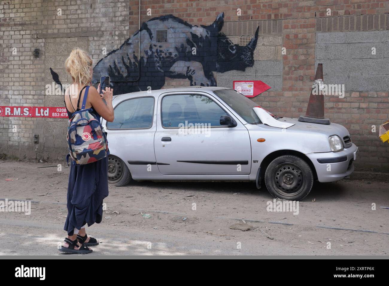 Une femme prend une photo d'une nouvelle œuvre d'art dévoilée par Banksy, représentant un rhinocéros qui semble grimper sur une voiture, la huitième œuvre d'art de sa collection sur le thème des animaux, sur Westmoor Street à Charlton, au sud-est de Londres. Date de la photo : lundi 12 août 2024. Banque D'Images