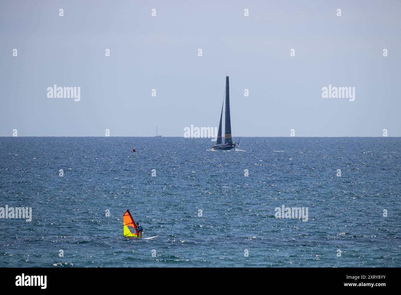 Barcelone, Espagne ; 2 juillet 2024 : Luna Rossa équipe italienne America's Cup AC75 glisse sur l'eau à grande vitesse pendant qu'un jeune essaie la planche à voile. Banque D'Images