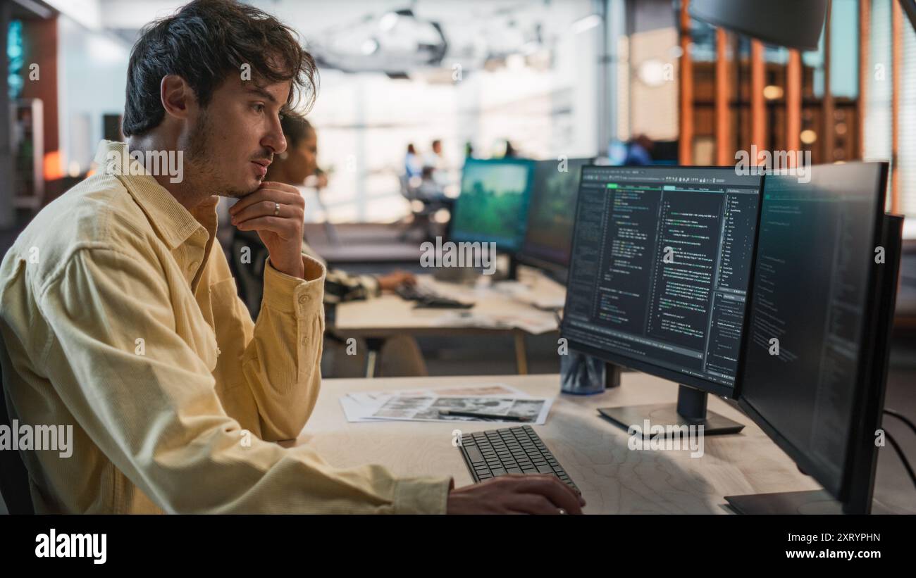 Programmeur de jeu caucasien masculin codage sur ordinateur de bureau dans Game Design Studio diverse Office. Focused Man écrit des lignes de code, fait de l'ingénierie de gameplay pour le nouveau jeu vidéo de survie immersive 3D. Banque D'Images