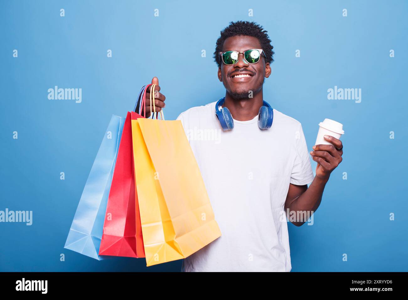 Portrait de jeune homme noir saisissant des sacs à provisions et tasse de papier de café à la main. Afro-américain portant des lunettes de soleil et souriant tout en portant des colis après avoir acheté des vêtements en vente. Banque D'Images